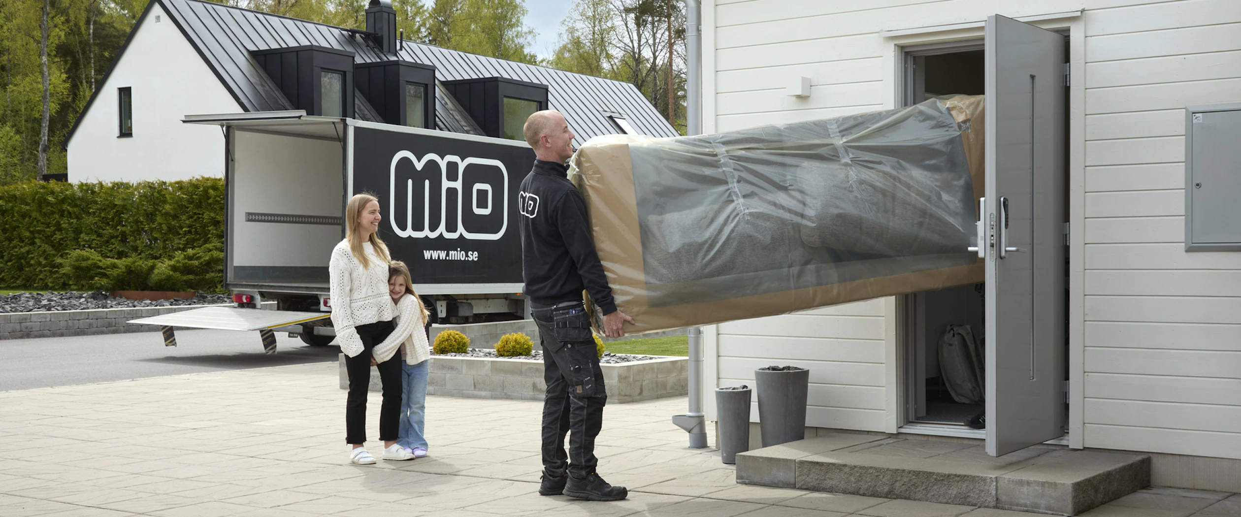 Mor och dotter ser på när medarbetare från Mio bär in en inplastad soffa in genom ytterdörren på en vit villa.