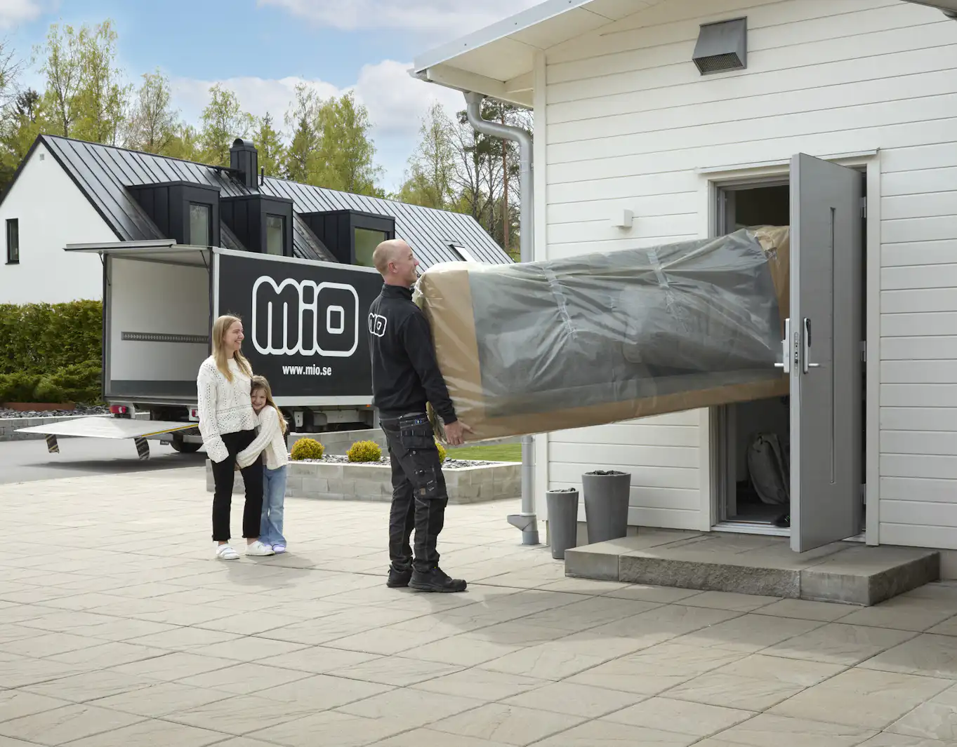 Mor och dotter ser på när medarbetare från Mio bär in en inplastad soffa in genom ytterdörren på en vit villa.