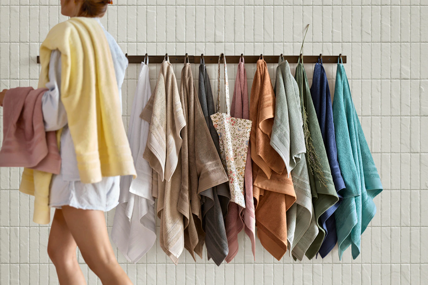 Kvinna i badrumsmiljö går förbi handdukskrokar med handdukar i olika färger. 
