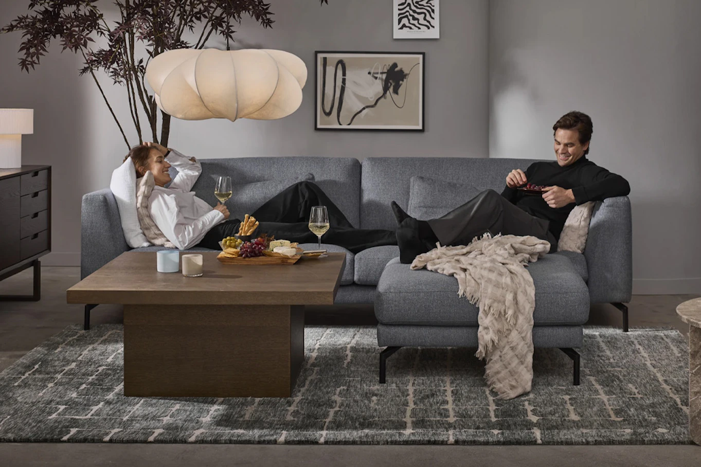 Kvinna ligger leendes i en grå soffa med vinglas i handen, en man sitter uppkrupen i motsatta hörnet.