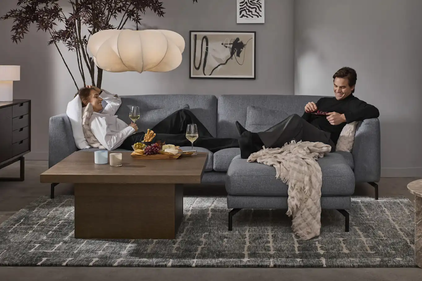 Kvinna ligger leendes i en grå soffa med vinglas i handen, en man sitter uppkrupen i motsatta hörnet.