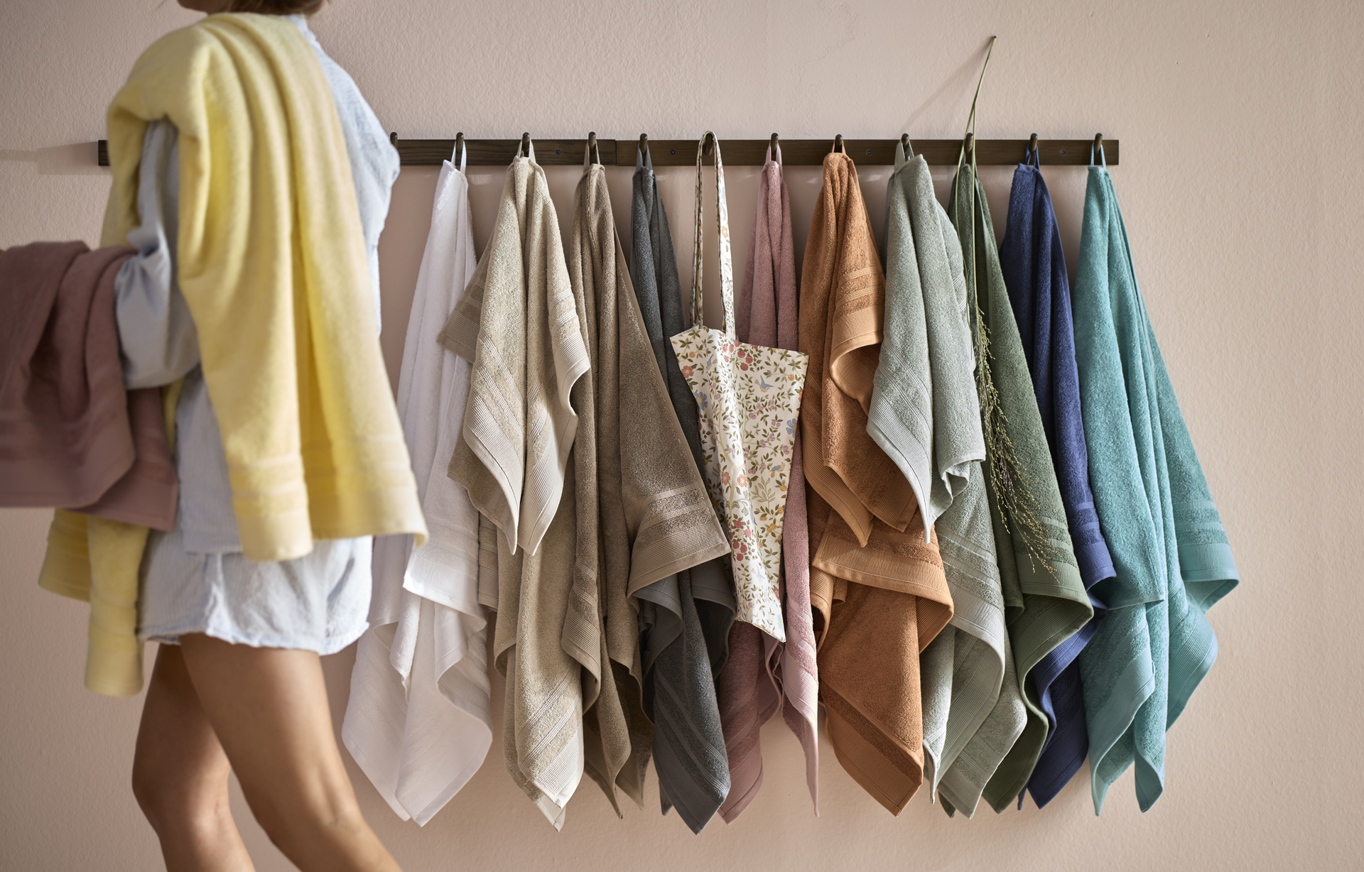 Kvinna i badrumsmiljö går förbi handdukskrokar med handdukar i olika färger.