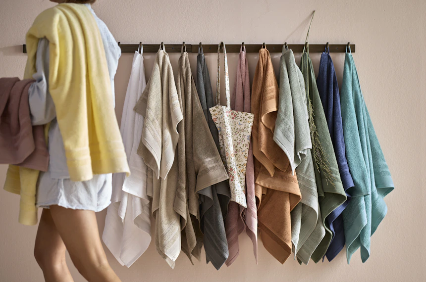 Kvinna i badrumsmiljö går förbi handdukskrokar med handdukar i olika färger.