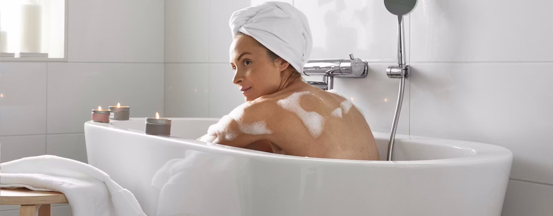 Kvinna med vit handduk virad om året sitter i badet med doftljus tända som står på kanten av badkaret.