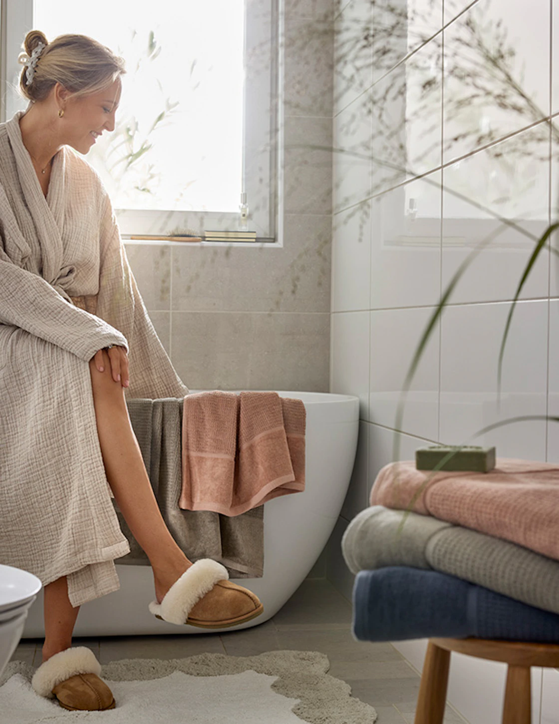 Kvinna sitter på badkarskanten i ett ljust badrum med solen som skiner in i fönstret, runtom finns handdukar i flera färger. 
