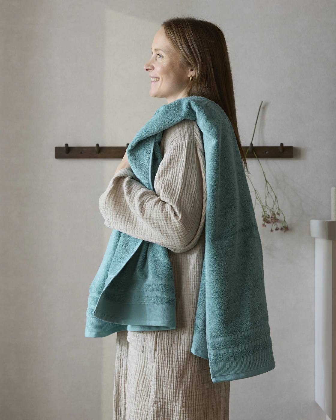 Kvinna iklädd morgonock står i badrum med en turkos handduk slängd över axeln. 