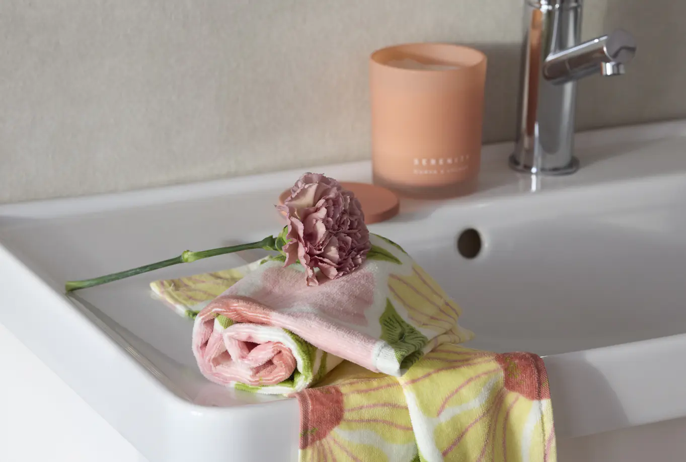 Blommig handduk ligger snett över ett handfat i badrum, en blomma ligger ovanpå handduken.