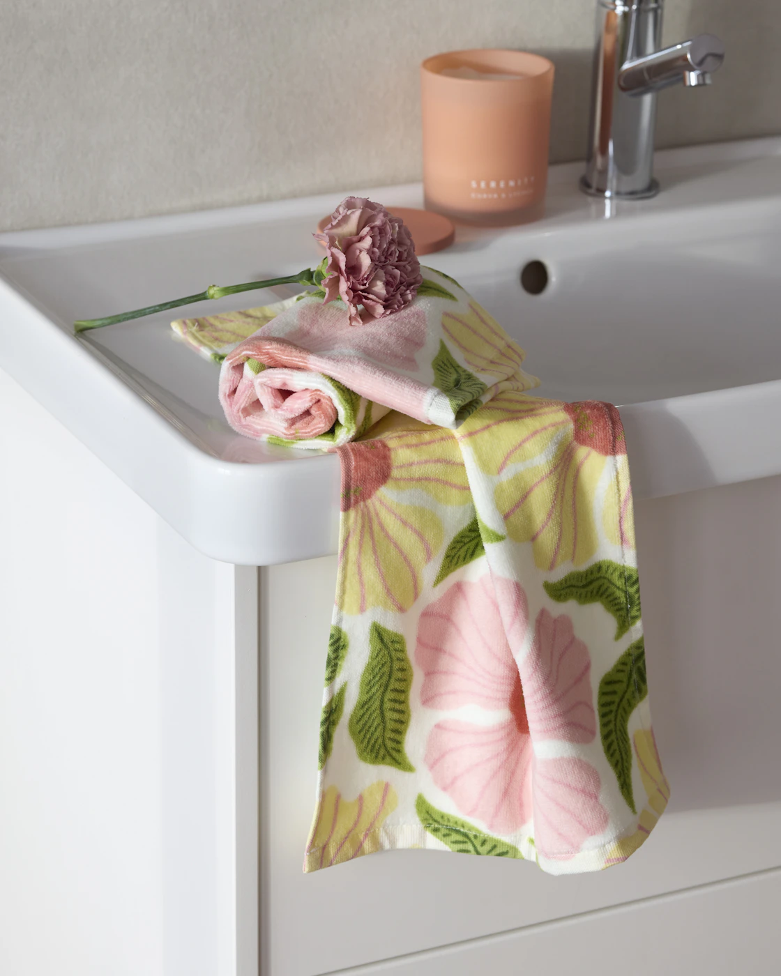 Blommig handduk ligger snett över ett handfat i badrum, en blomma ligger ovanpå handduken. 