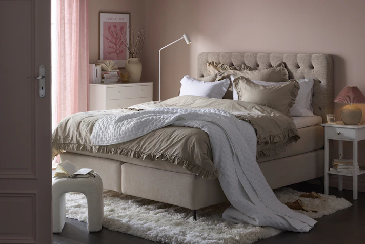 en beige kontinentalsäng står i ett sovrum med ljusrosa väggar och en vit matta