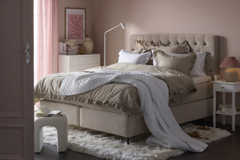 en beige kontinentalsäng står i ett sovrum med ljusrosa väggar och en vit matta