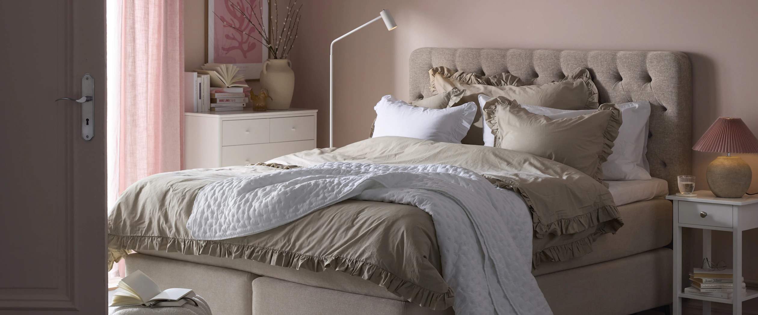 en beige kontinentalsäng står i ett sovrum med ljusrosa väggar och en vit matta