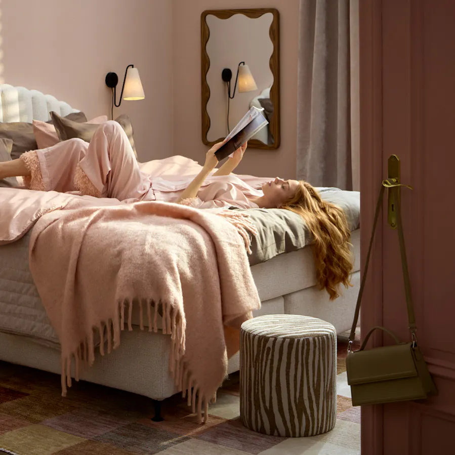 Kvinna ligger ledigt på rygg i kontinentalsäng och läser en bok med ena benet över det andra. 