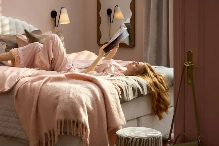 Kvinna ligger ledigt på rygg i kontinentalsäng och läser en bok med ena benet över det andra.