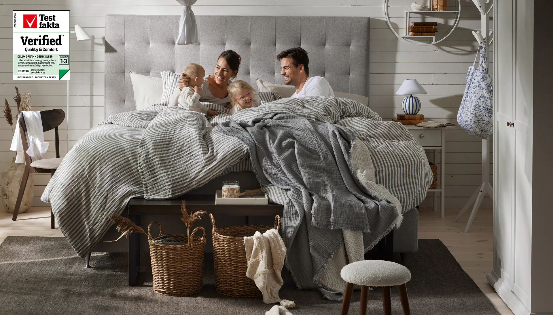 Barnfamilj med kvinna, man och två små barn ligger i kontinentalsäng och skrattar tillsammans. 