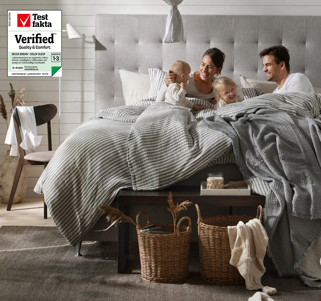 Barnfamilj med kvinna, man och två små barn ligger i kontinentalsäng och skrattar tillsammans.