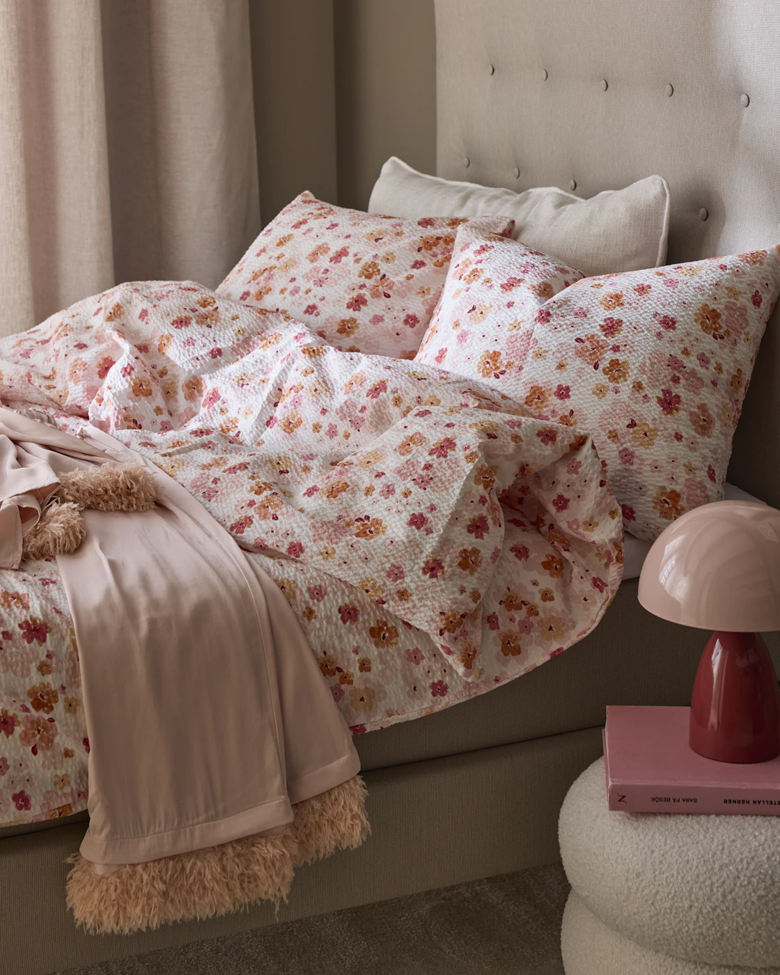 En säng är bäddad med ett påslakanset med rosa blommor i bäckebölja, på nattduksbordet står en rosa och röd lampa