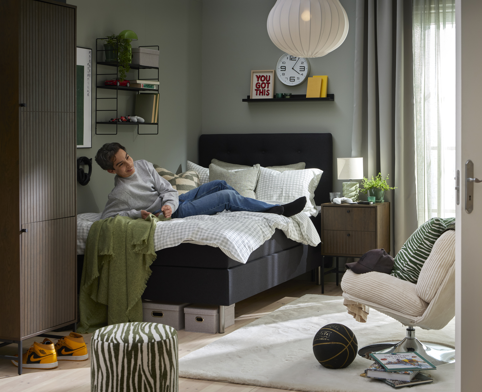 Ungdom ligger ned i säng klädd i mörkgrått tyg i ett ungdomligt inspirerat sovrum. 