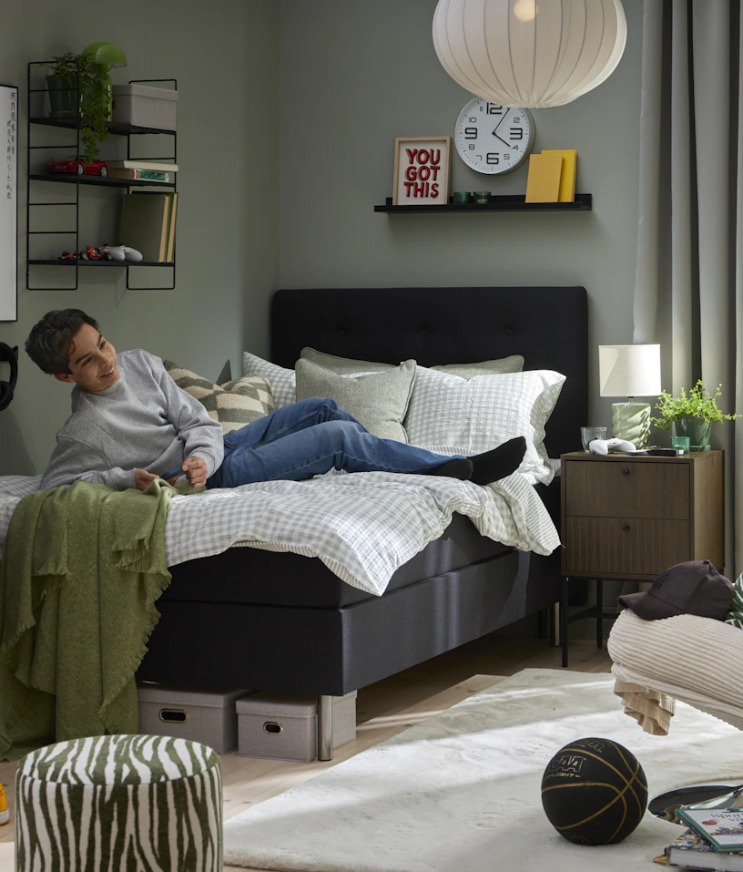 Ungdom ligger ned i säng klädd i mörkgrått tyg i ett ungdomligt inspirerat sovrum.