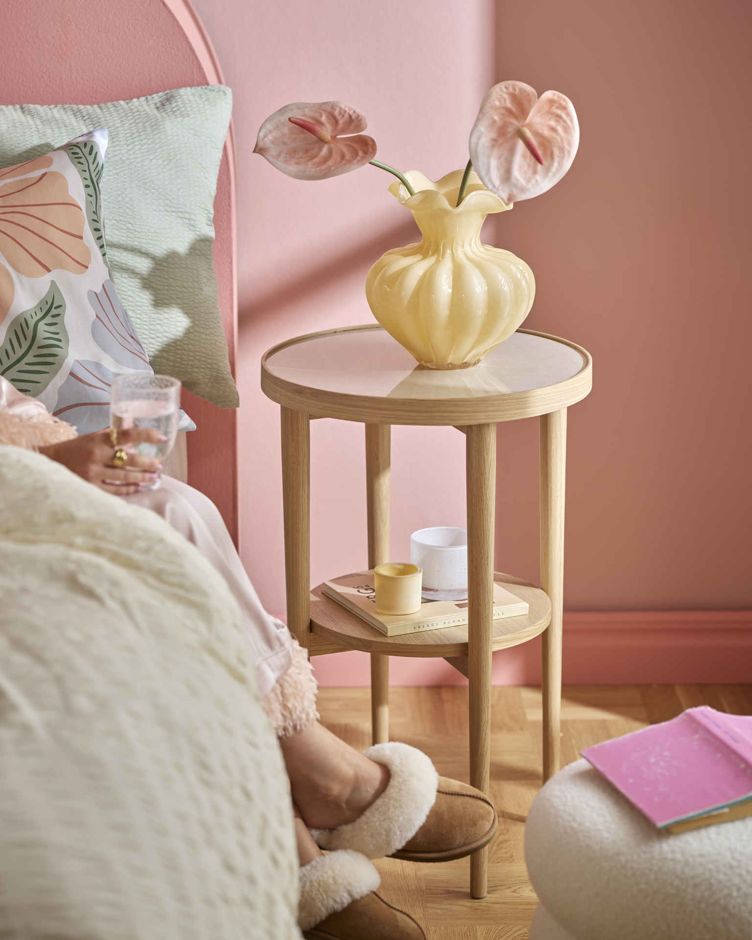 I ett rosa sovrum står ett sängbord i ljust trä med en gul vas och en rosa snittblomma.