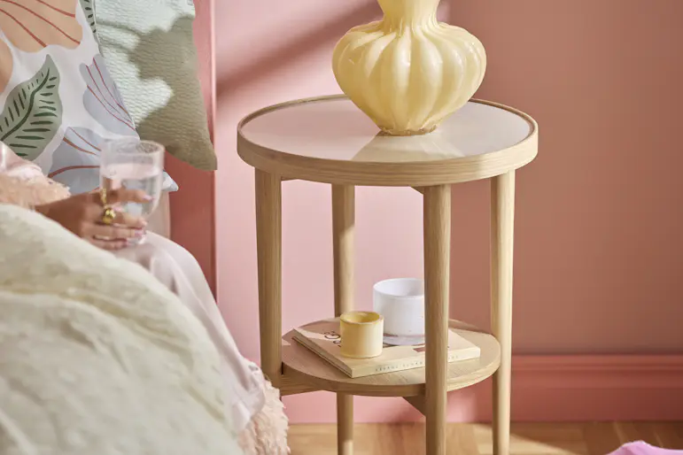 Ett sängbord i ek står bredvid en säng i ett rosa sovrum. På sängbordet står en gul vas med rosa blommor.