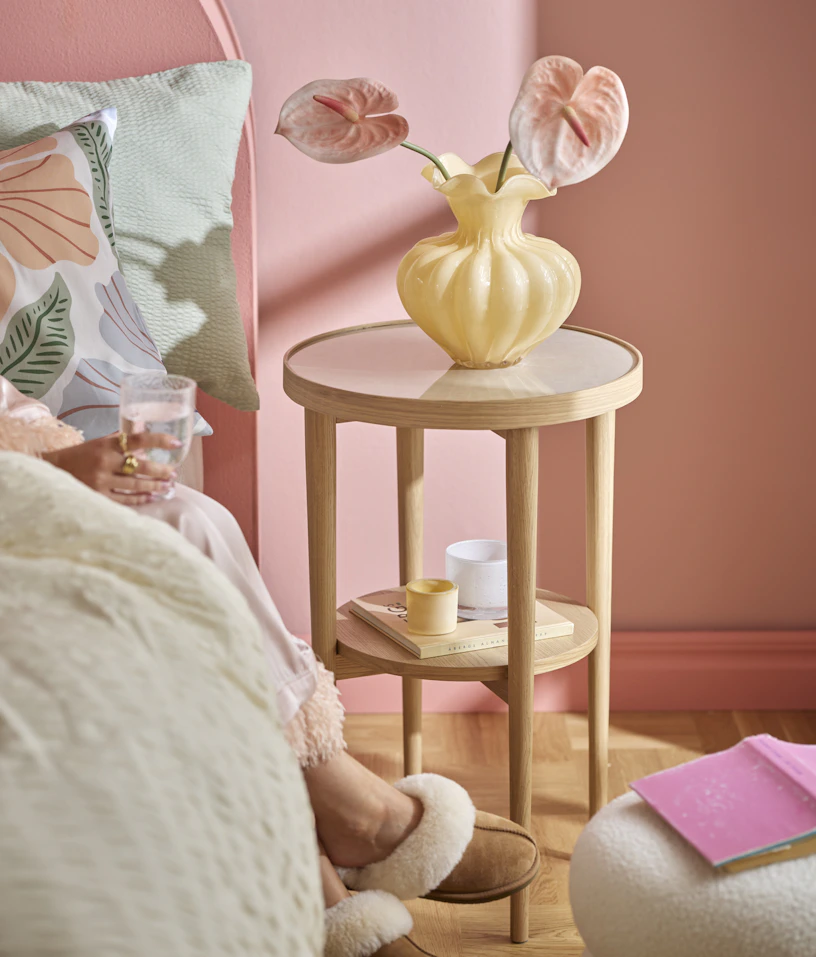 I ett rosa sovrum står ett sängbord i ljust trä med en gul vas och en rosa snittblomma.