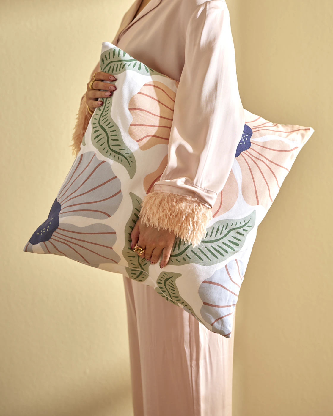 En kvinna står i ett gult rum och håller en kudde med storblommigt mönster under armen