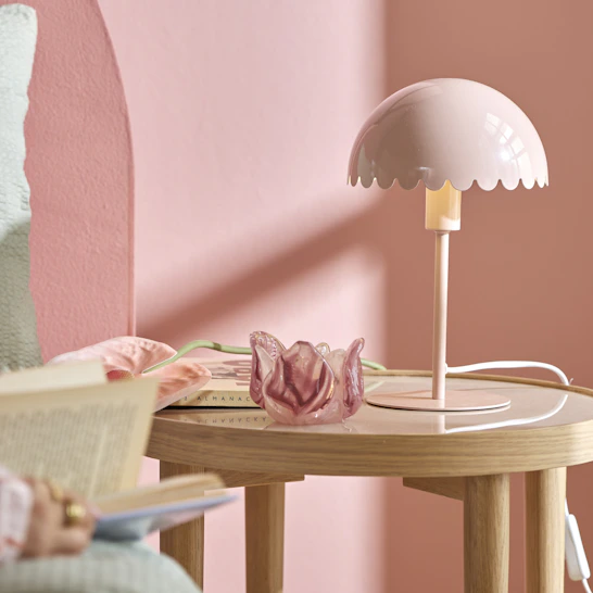 I ett rosa sovrum står ett sängbord i ljust trä, en rosa bordslampa och en rosa ljuslykta.