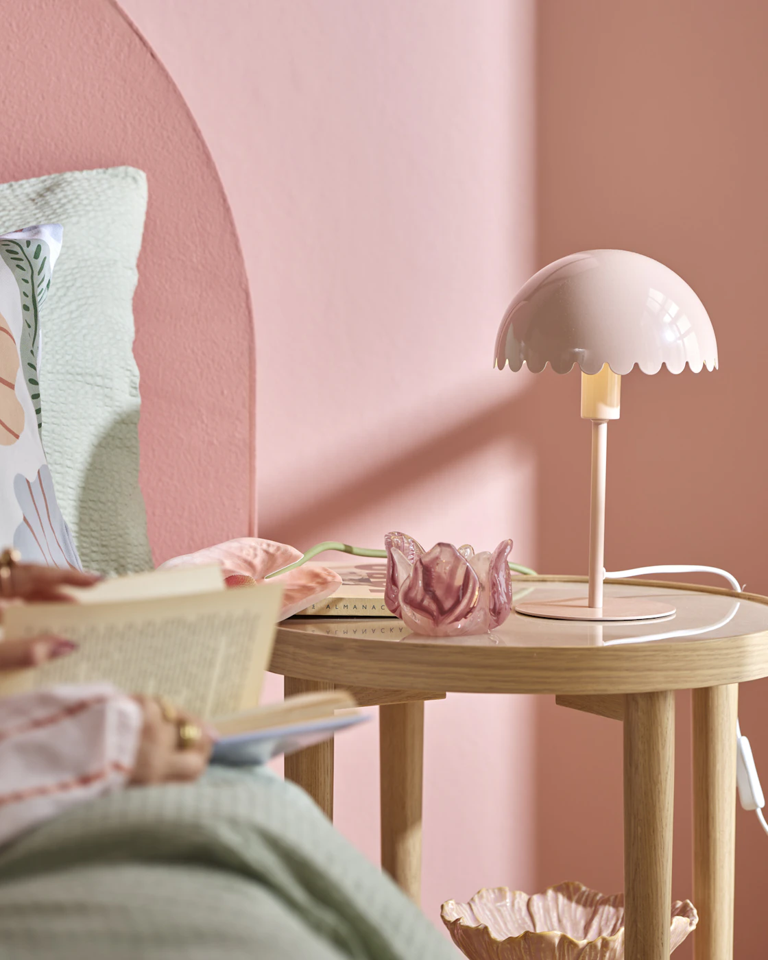 På ett nattduksbord står en rosa lampa bredvid en ljusstake i rosa glas