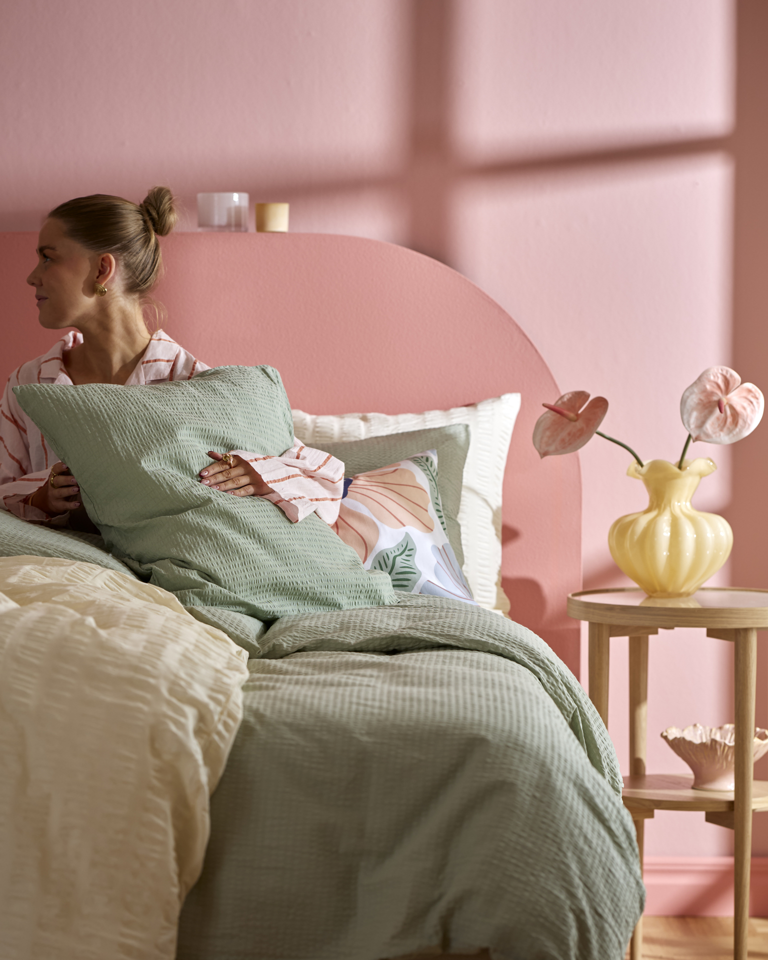 I ett rosa sovrum sitter en kvinna i en säng bäddad med påslakan i gult och grönt