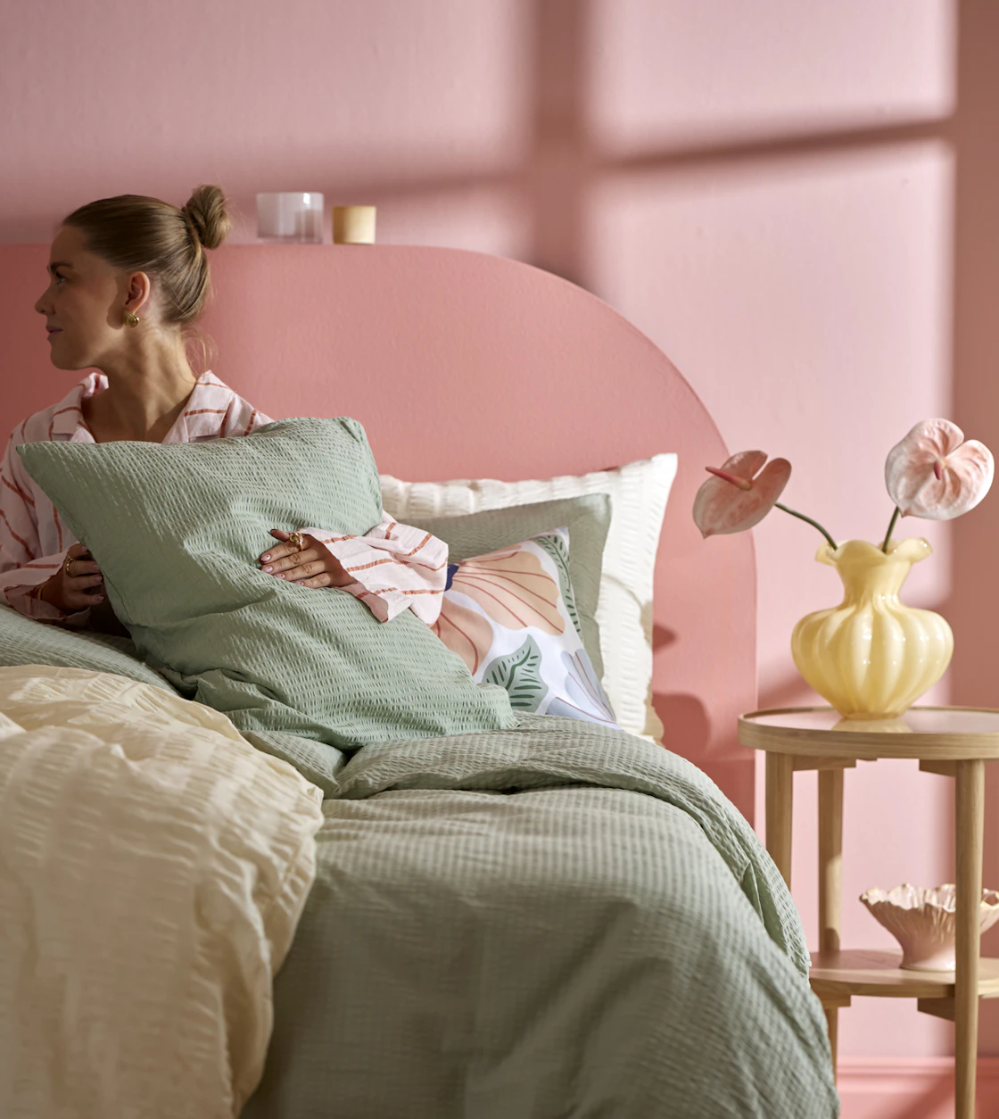 I ett rosa sovrum sitter en kvinna i en säng bäddad med påslakan i gult och grönt