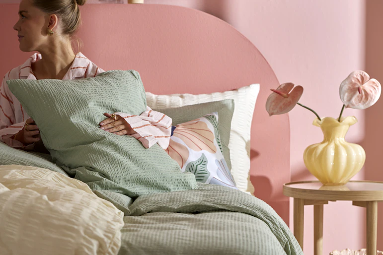 En kvinna ligger i en säng som är bäddad med gröna och gula sängkläder i bäckebölja.