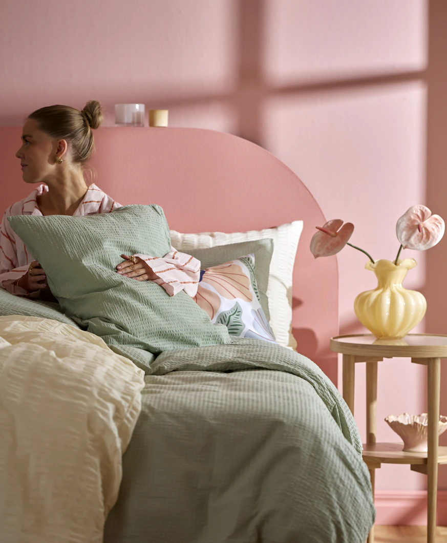 En kvinna ligger i en säng som är bäddad med gröna och gula sängkläder i bäckebölja.