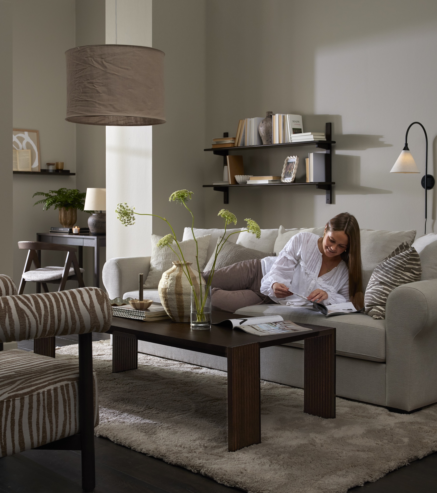 I ett grått vardagsrum ligger en kvinna i en ljusgrå soffa och bläddrar i ett magasin.