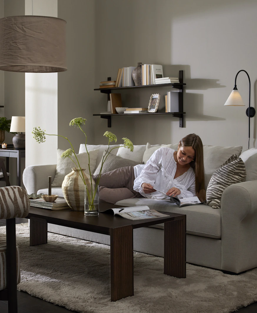 I ett grått vardagsrum ligger en kvinna i en ljusgrå soffa och bläddrar i ett magasin.