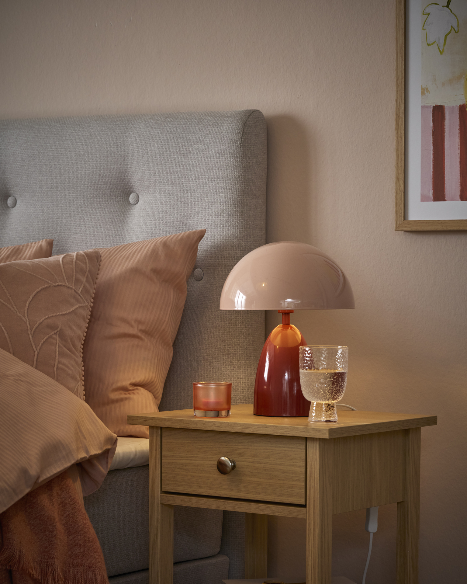 En rosa lampa med röd fot står på ett sängbord i ljust trä.