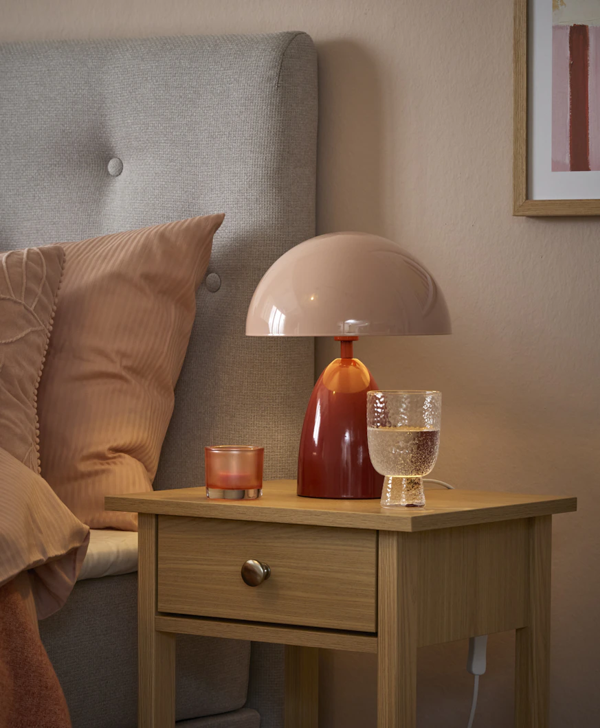 En rosa lampa med röd fot står på ett sängbord i ljust trä.
