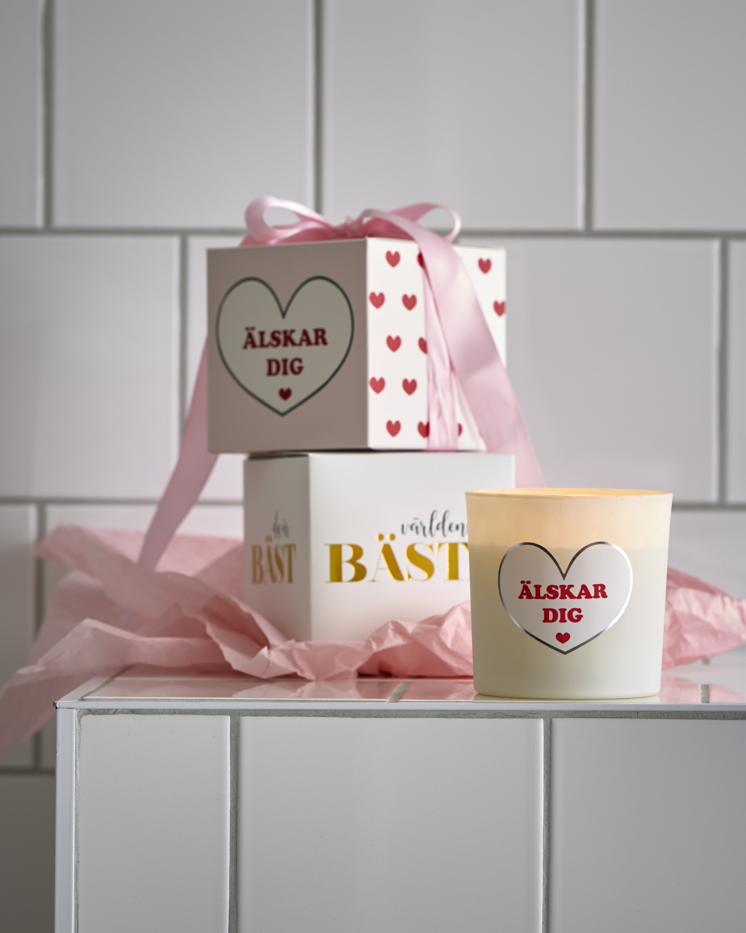 Doftljus med budskapen "Älskar dig" och "Världens bästa" har en rosa rosett om sig och står framför en kaklad vägg.