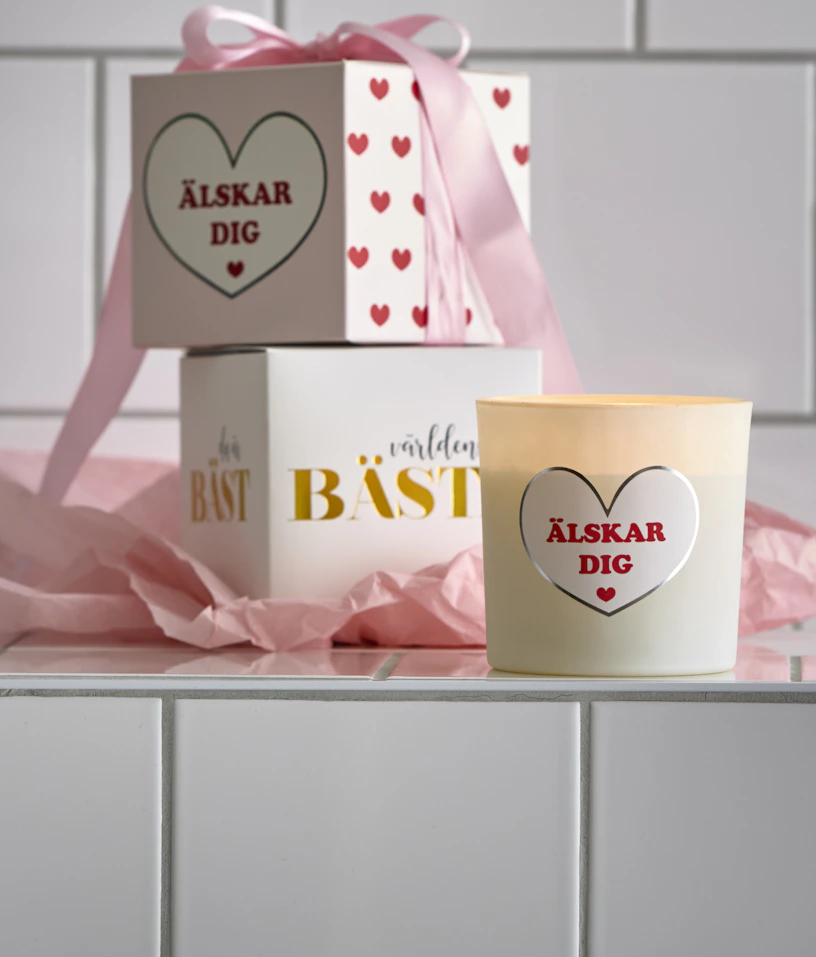 Doftljus med budskapen "Älskar dig" och "Världens bästa" har en rosa rosett om sig och står framför en kaklad vägg.