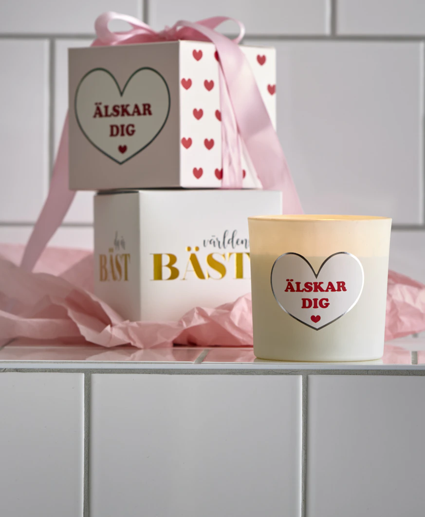 Doftljus med budskapen "Älskar dig" och "Världens bästa" har en rosa rosett om sig och står framför en kaklad vägg.