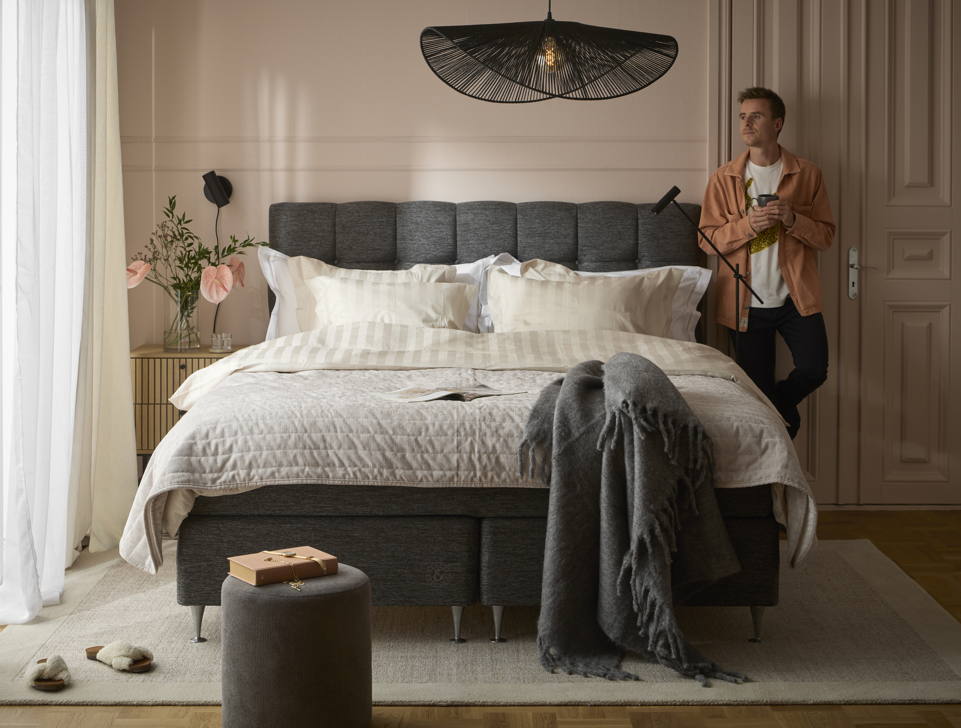 Ung man med skjorta står lutandes med ryggen mot väggen intill sänggavel och blickar ut mot fönster i sovrum. 