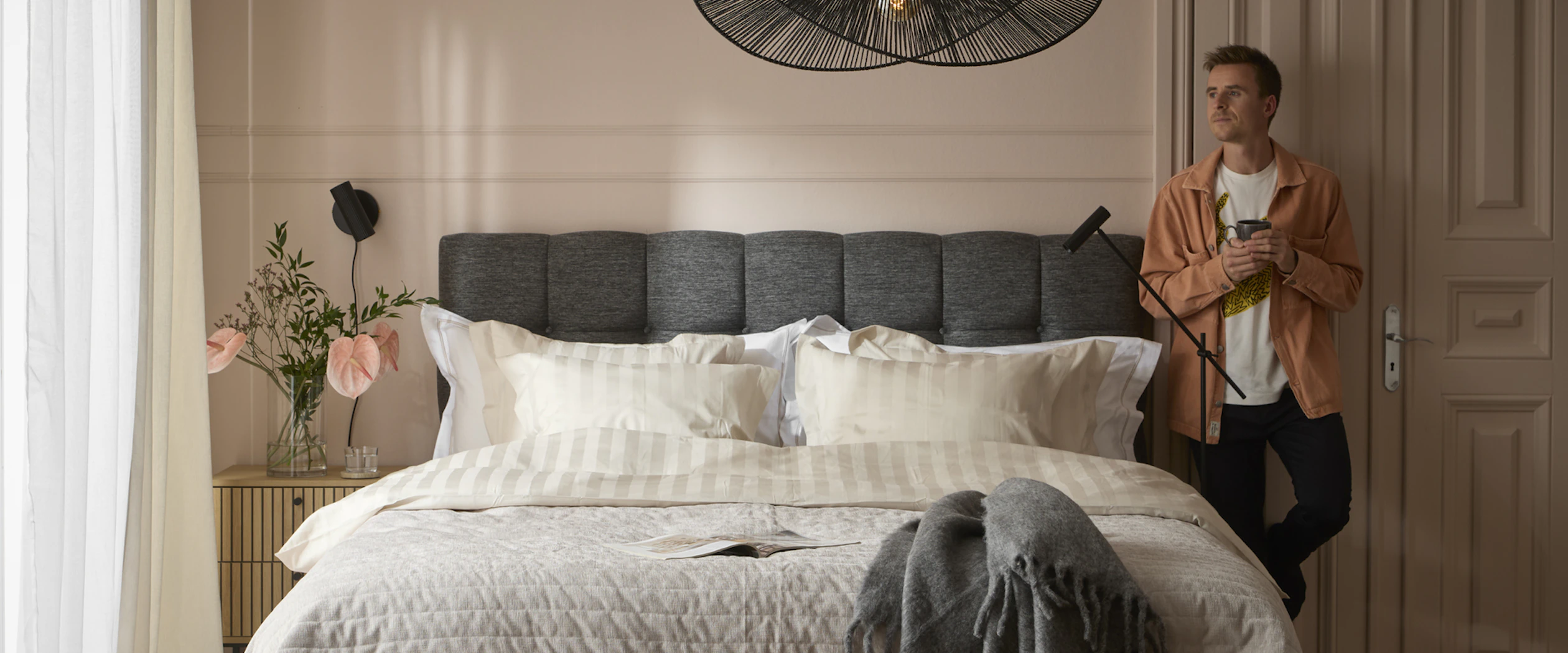 Ung man med skjorta står lutandes med ryggen mot väggen intill sänggavel och blickar ut mot fönster i sovrum.