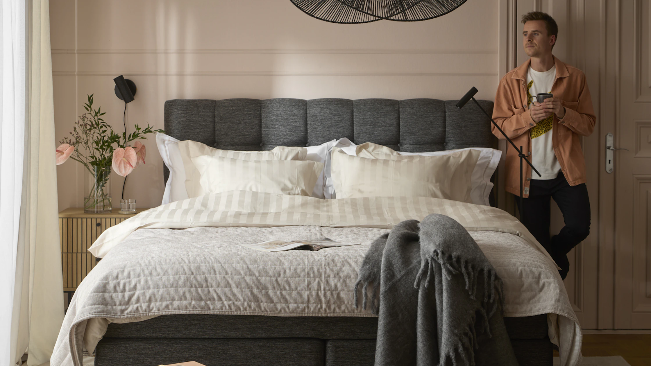 Ung man med skjorta står lutandes med ryggen mot väggen intill sänggavel och blickar ut mot fönster i sovrum.