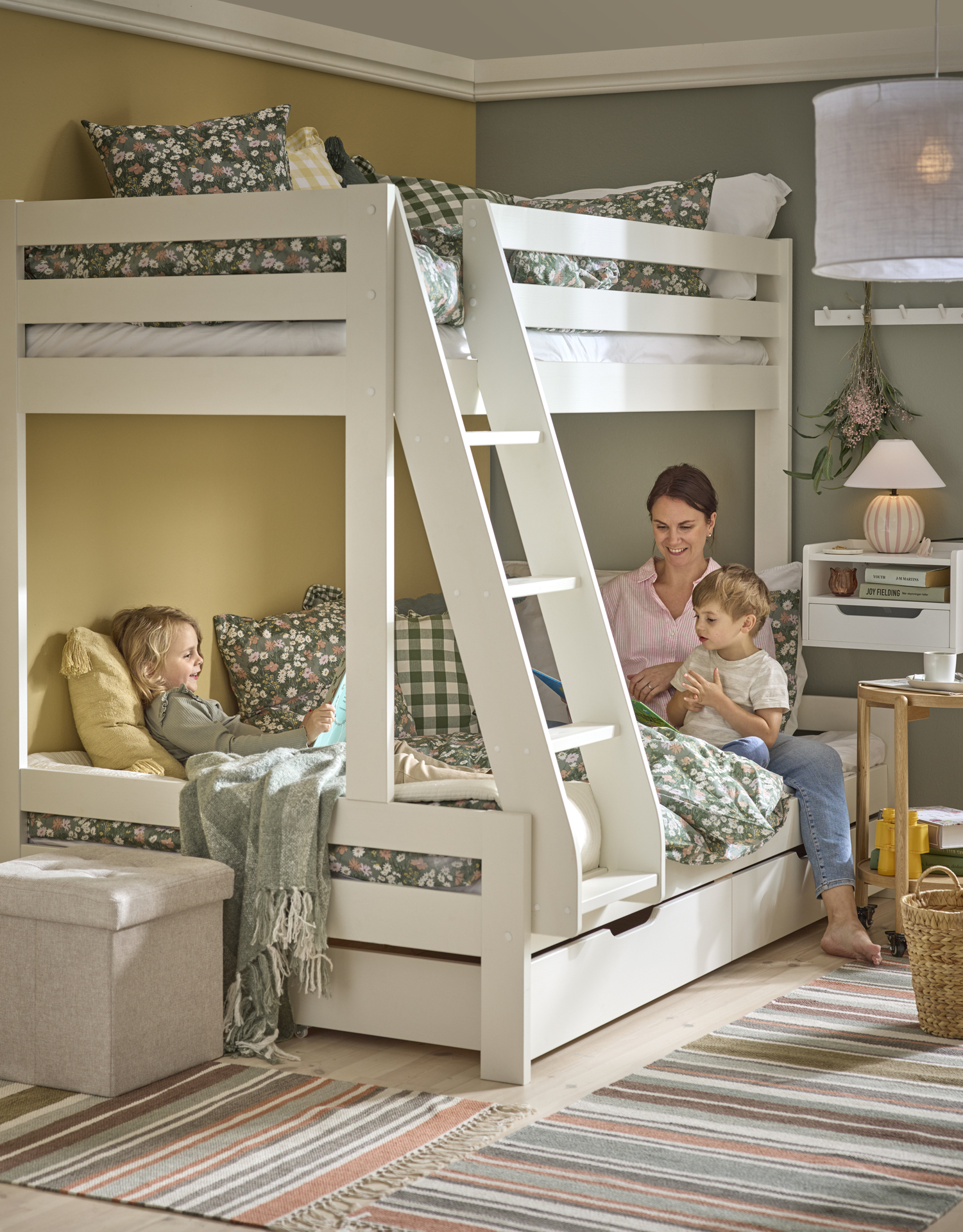 En kvinna och två barn läser bok i en våningssäng bäddad med grönblommiga sängkkäder.