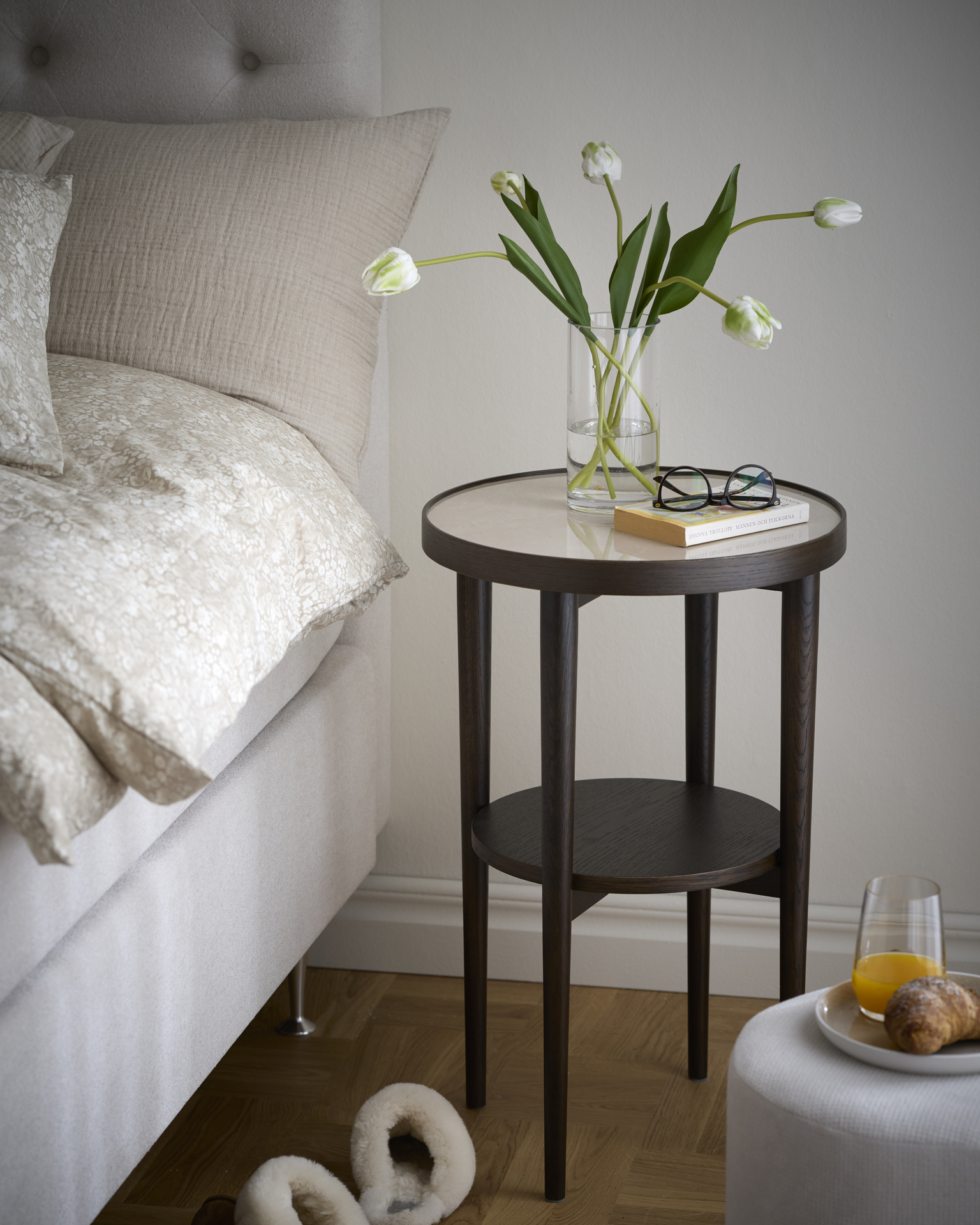 En vas med vita tulpaner, en bok och ett par glasögon står på ett sängbord i mörkt trä med keramikskiva.