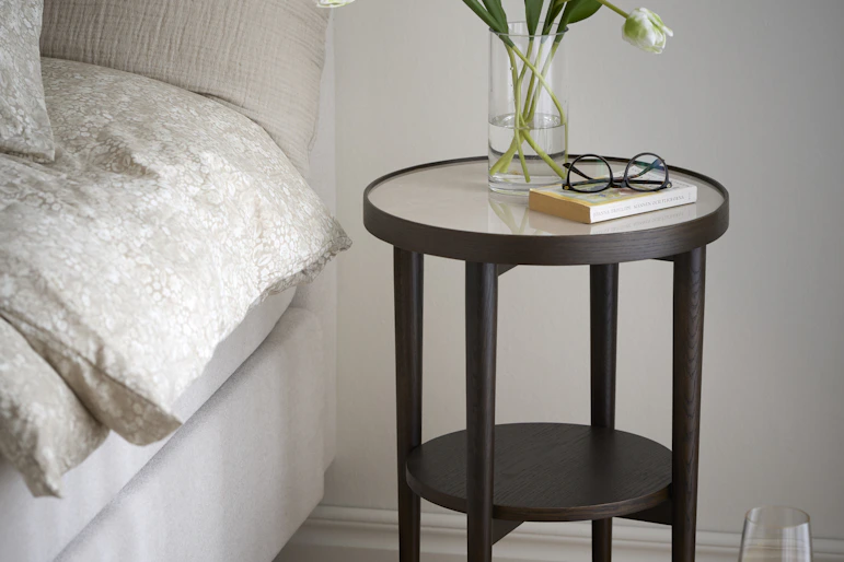 En vas med vita tulpaner, en bok och ett par glasögon står på ett sängbord i mörkt trä med keramikskiva.