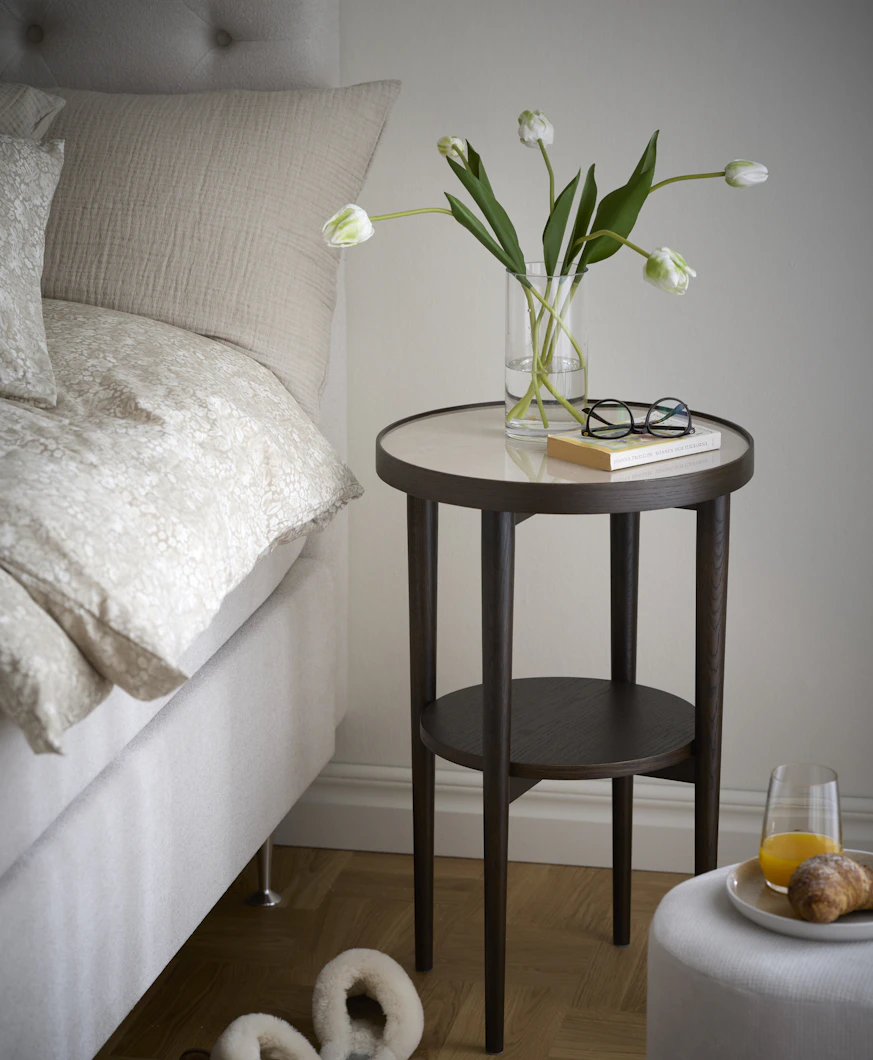 En vas med vita tulpaner, en bok och ett par glasögon står på ett sängbord i mörkt trä med keramikskiva.