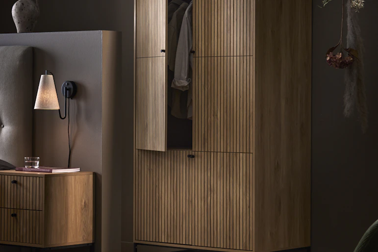 En garderob i mörkt trä med ribbor och matchande sängbord står i ett mörkt sovrum.