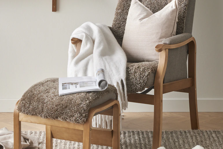 I en brun fårskinnsfåtölj med fotpall ligger en vit kudde, en vit pläd och ett uppslaget magasin.