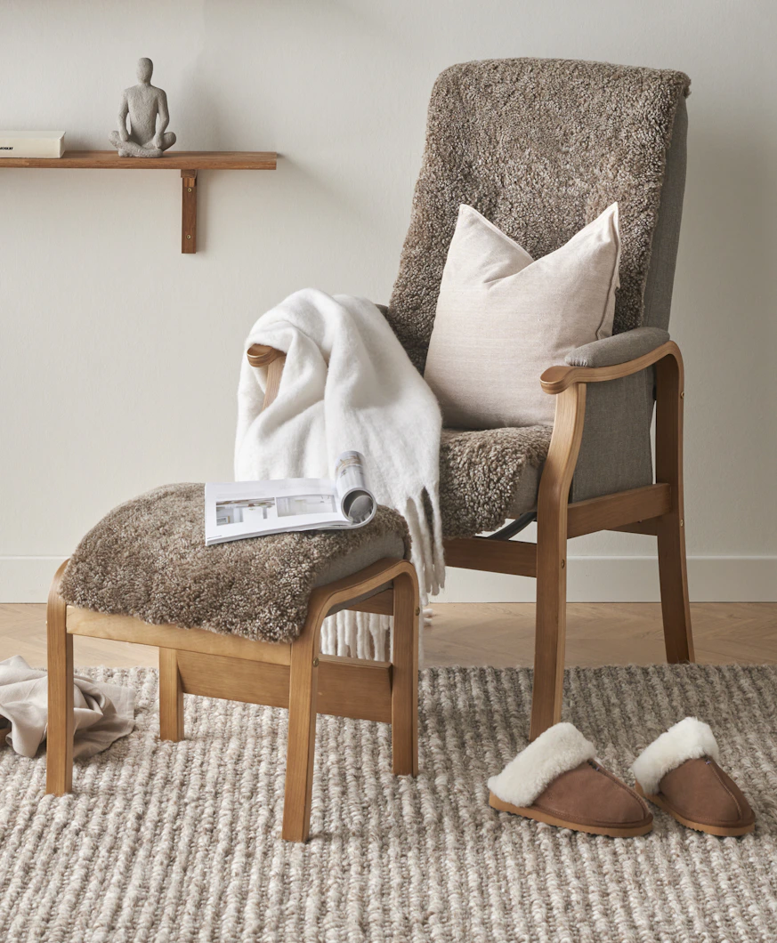 I en brun fårskinnsfåtölj med fotpall ligger en vit kudde, en vit pläd och ett uppslaget magasin.