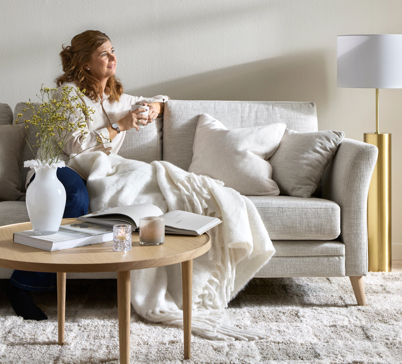 I ett avskalat vardagsrum sitter en kvinna med en kopp te i en ljusgrå soffa